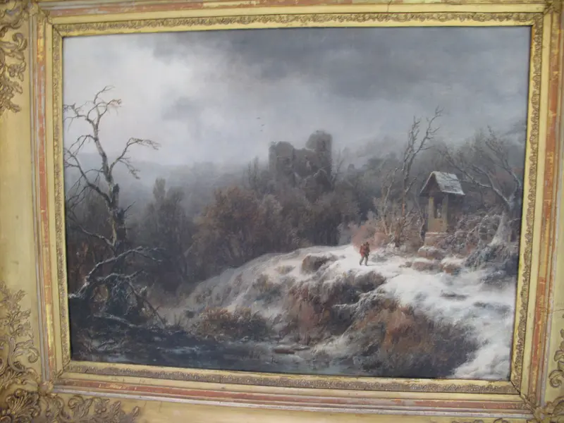 A solitary figure walks along a snow-covered path framed by bare trees and ancient ruins under a heavy winter sky.