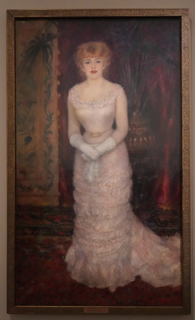 A young woman in a pale pink ruffled gown with gloved hands crossed at her waist stands against a shadowy background with decorative patterns and drapery.
