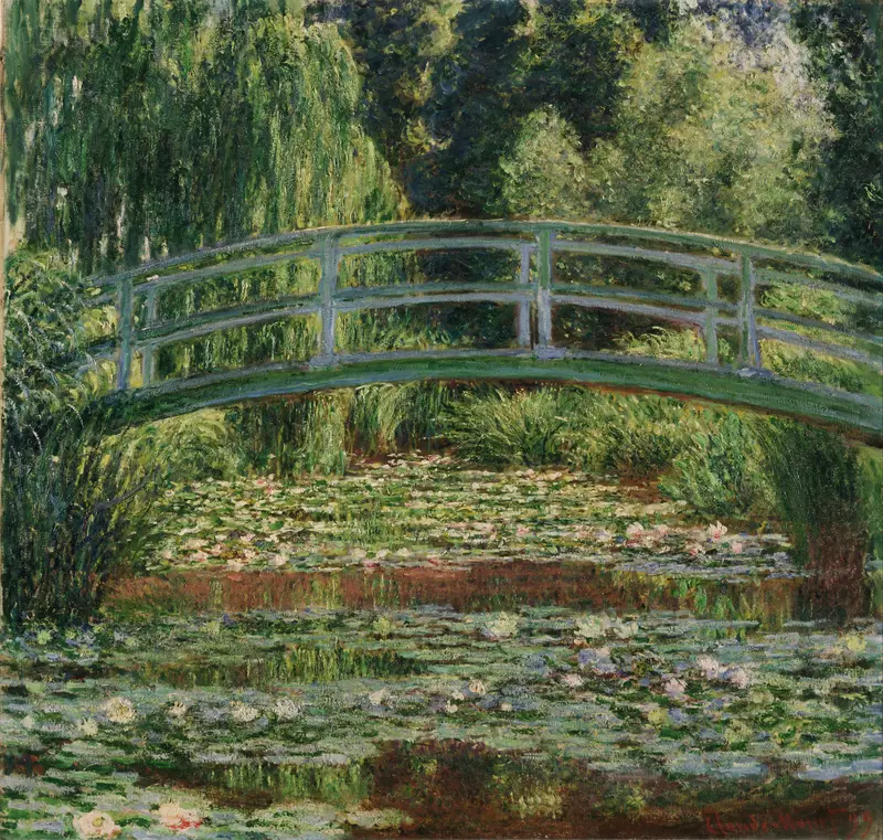A curved wooden bridge arches over a pond densely covered with pale pink and white water lilies, surrounded by green foliage and reflected in rippling water.