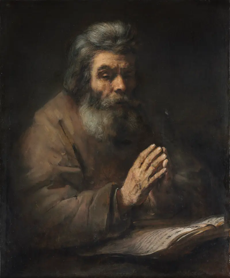 A bearded elderly man in a heavy cloak sits with hands pressed together in prayer before an open book, illuminated against a dark background.