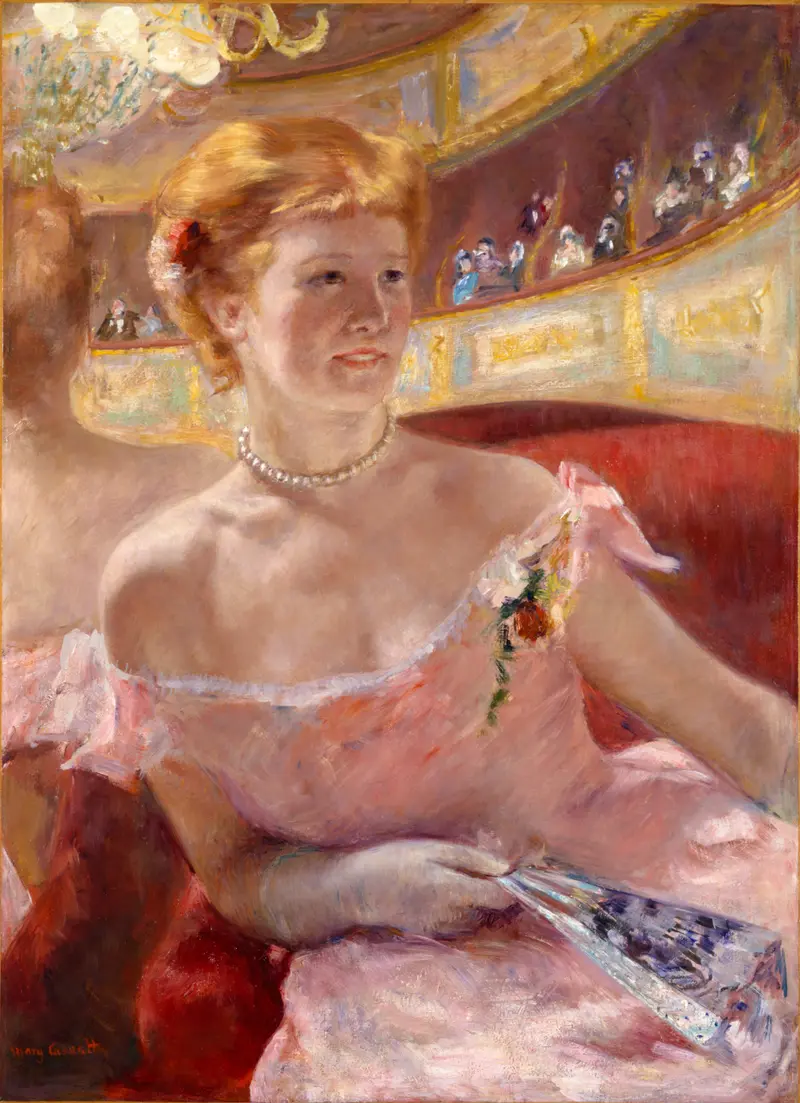 A woman in a peach dress with a pearl necklace sits in a theater box holding a blue and white fan, with a mirror reflecting the ornate interior behind her.
