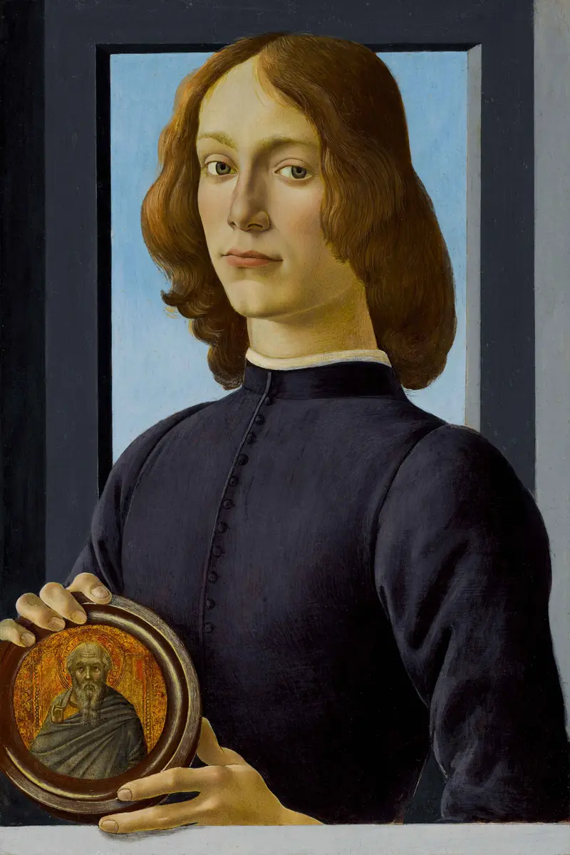 A young man with long wavy hair holds a circular medallion showing a bearded saint, standing before a window with a calm expression.