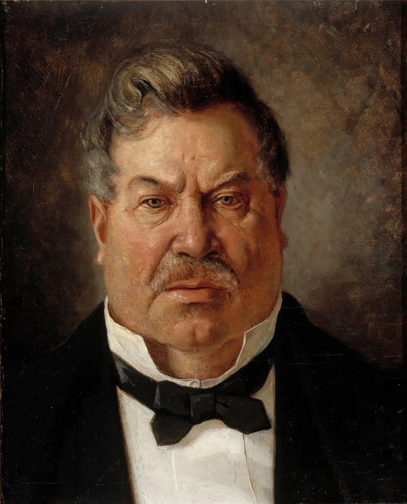 A man with grey wavy hair and a ruddy face wears a black jacket, white shirt, and black bow tie against a warm brown and grey background.