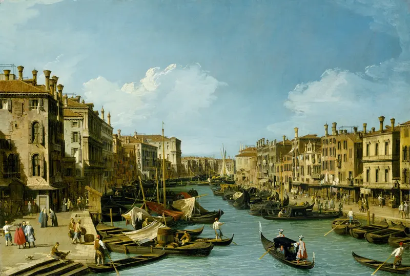 A broad view of the Grand Canal near the Rialto Bridge in Venice showing gondolas, boats, Venetian palazzi, and people gathered along the stone embankment under a blue sky.