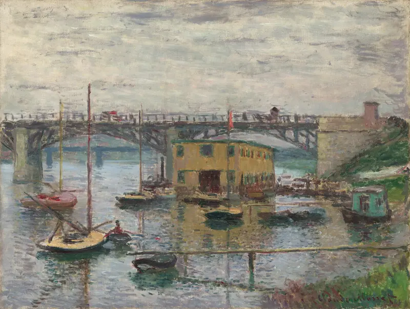 An iron bridge spans across a gray sky above calm water with several boats, a yellow building, and a steep riverbank in the background.