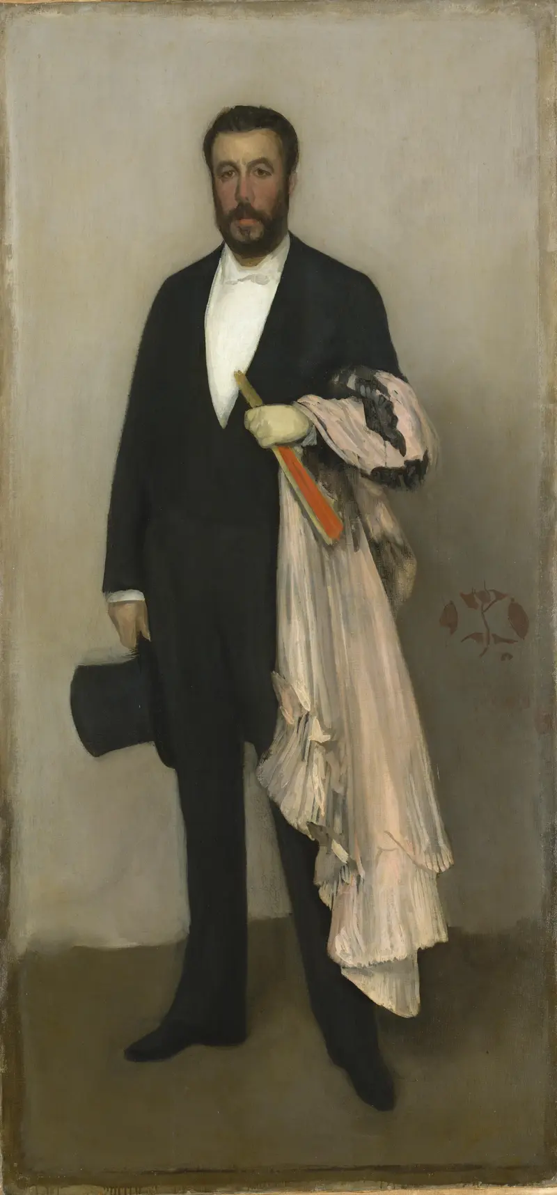 A man in formal black attire stands upright, holding a delicate garment and fan against a neutral background.