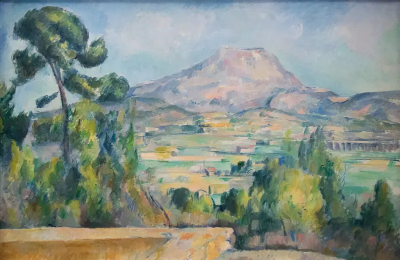 A broad mountain with pale slopes rises behind cultivated fields, scattered buildings, a tall pine tree, and dense clusters of trees under a luminous blue sky.