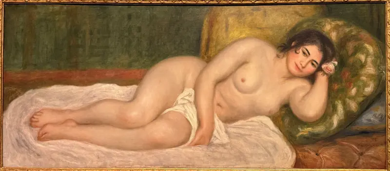 A nude woman reclines on a white sheet, her head resting on a green and yellow floral cushion, with a pink and white flower behind her ear.