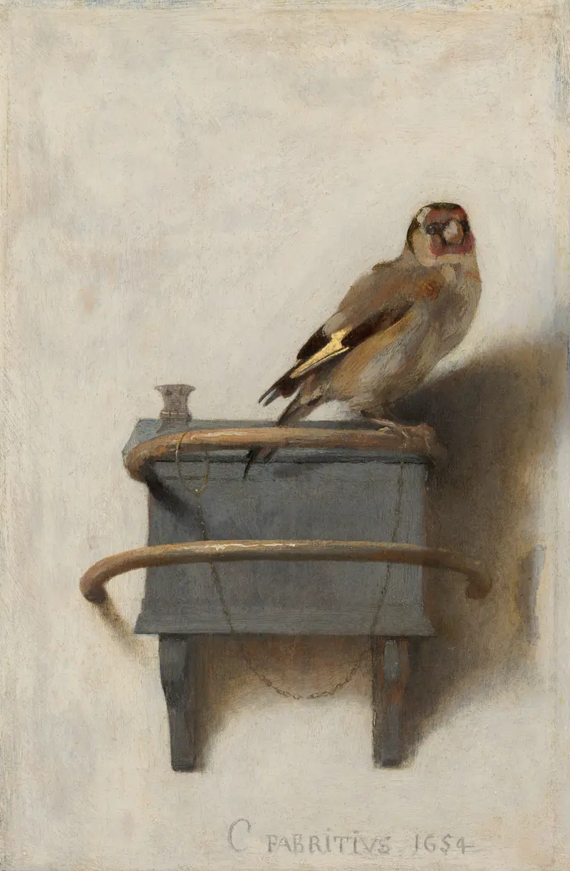 A European goldfinch with a red face mask and yellow wing stripe perches on a blue-grey box against a pale wall, chained by its ankle.